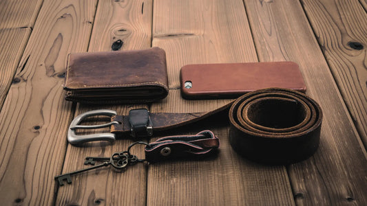 Rustic leather accessories including a wallet, belt, keychain, and phone case arranged on a wooden surface, showcasing handmade leather craftsmanship.