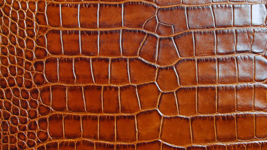 A close-up of a brown leather surface with a distinctive alligator or crocodile pattern. The textured leather displays a glossy finish, highlighting the intricate details of the scales and the natural variations in the design. 