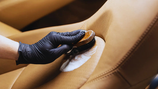 A close-up of a person wearing a black glove, cleaning a tan leather surface with a soft-bristle brush and leather cleaning solution. The person is gently scrubbing, creating a foamy lather that helps to remove dirt and maintain the leather's condition. 