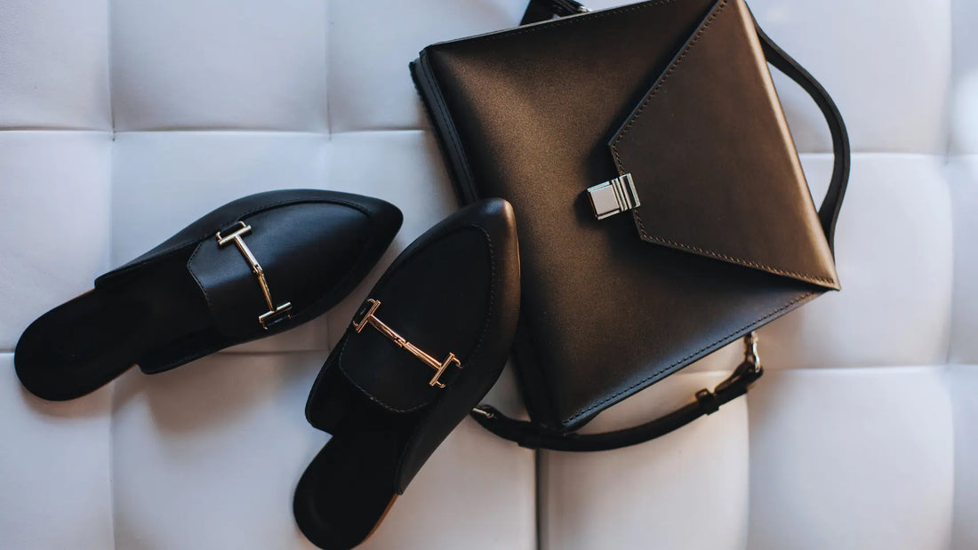 A stylish arrangement of elegant black leather accessories placed on a white, tufted surface. The items include a pair of sleek leather loafers with gold metal hardware and a structured leather handbag with a metallic clasp.